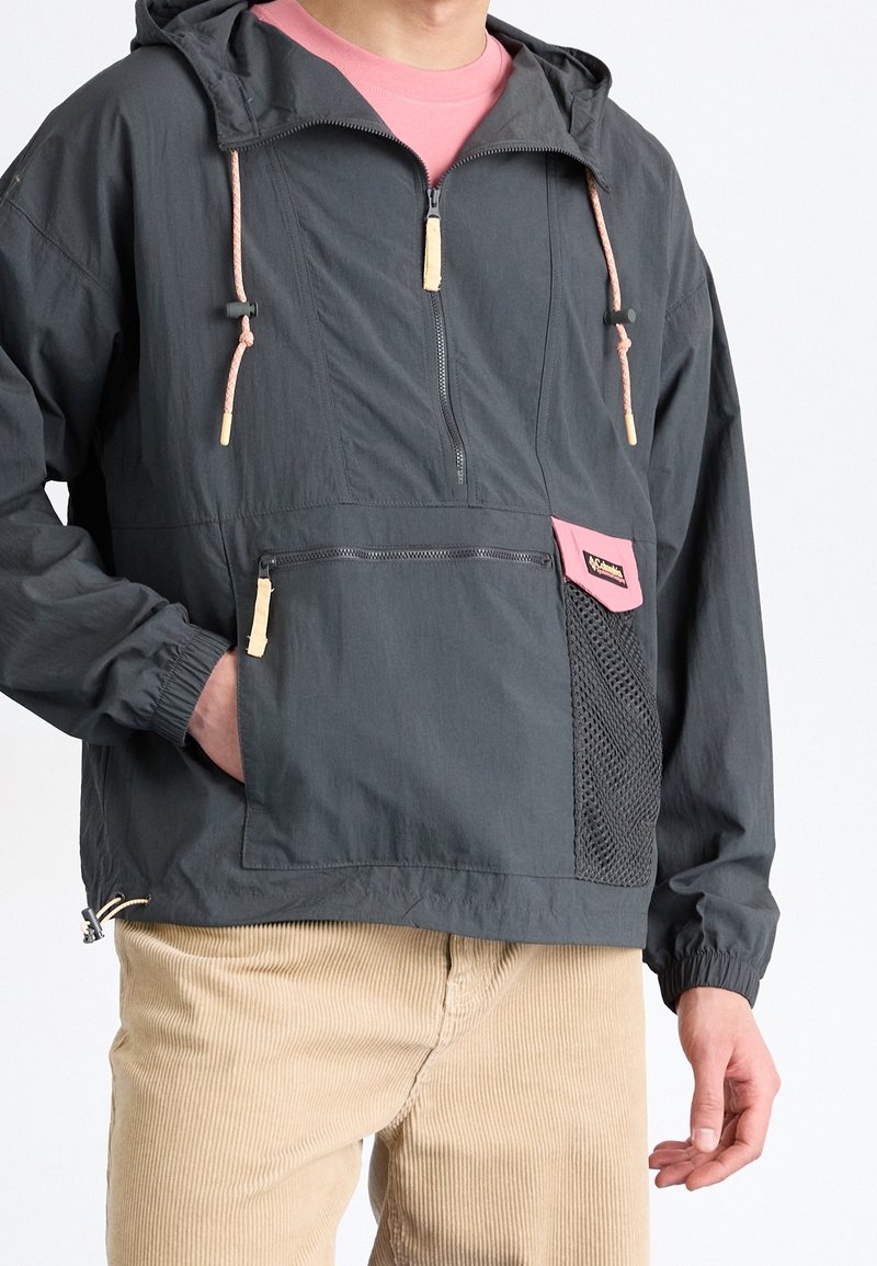 Personne portant une veste à capuche gris foncé avec cordons roses et écusson, un pantalon beige et une chemise rose en dessous.