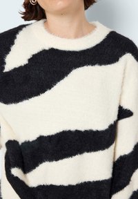 Kremowy sweter z wyrazistymi, nieregularnymi czarnymi paskami. Wykonany z miękkiego, teksturowanego materiału, z okrągłym dekoltem i prążkowanym wykończeniem.