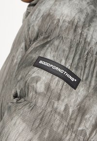 Grå puffjacka med en texturerad yta. Har en svart etikett på ärmen som har texten "GOODFORNOTHING" i vita bokstäver.