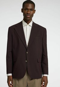 Brązowa marynarka z dwoma guzikami, klapami i jedną kieszenią na piersi, noszona na paskowanej koszuli. Gładka tekstura i dopasowany krój.
