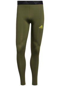 Olive zielone legginsy wykonane z elastycznego materiału, z czarnym pasem w talii z logo Adidas oraz małym żółtym logo na nodze.