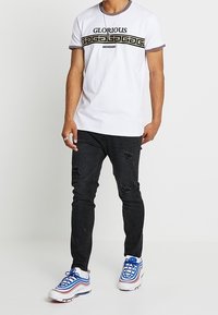 Camiseta blanca de manga corta con texto "GLORIOUS" en negro y dorado y banda con patrón. Pantalones vaqueros ajustados negros rasgados y zapatillas azules y blancas.