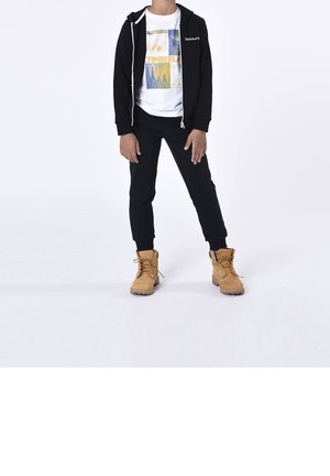 Barn iført sort lynlås-hættetrøje, hvid T-shirt med grafik, sorte bukser og beige snørebåndsstøvler står mod hvid baggrund.