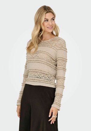 Femme blonde portant un top long à manches en dentelle beige avec des motifs floraux et une jupe noire, souriante et regardant vers la droite.