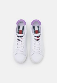 Sneaker bianchi in pelle con accenti viola sul colletto, lacci piatti e una soletta brandizzata con strisce rosse, bianche e blu.