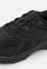 Chaussure de sport noire avec une tige en maille texturée, semelle en caoutchouc et détails renforcés. Caractérisée par des lacets plats et un col rembourré.