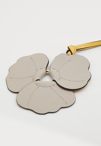 Le porte-clés en cuir présente trois composants de forme pétale beige avec des bords noirs contrastants et une sangle jaune. Les détails de couture visibles rehaussent le design.