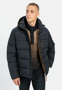 Schwarze Steppjacke mit Kapuze, ausgestattet mit einem gesteppten Design, seitlichen Taschen und braunen Akzenten. Wird über einem strukturierten Strickpullover getragen.