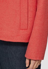 Koraalwollen jas met een zachte textuur, voorzien van een strakke stijl en een subtiele zakdetail aan de zijkant. Gestyled met donkerdenim.