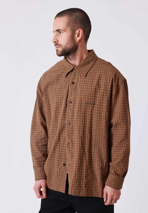 Homme aux cheveux courts et à la barbe portant une chemise marron à carreaux à manches longues boutonnée, regardant vers sa gauche sur un fond uni.