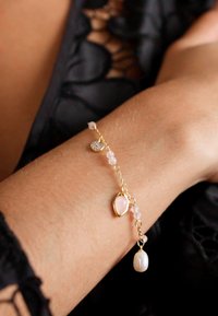 Gold bracelet featuring pink beads, a teardrop stone, and a pearl charm, showcasing a delicate and layered design on a skin-toned wrist.