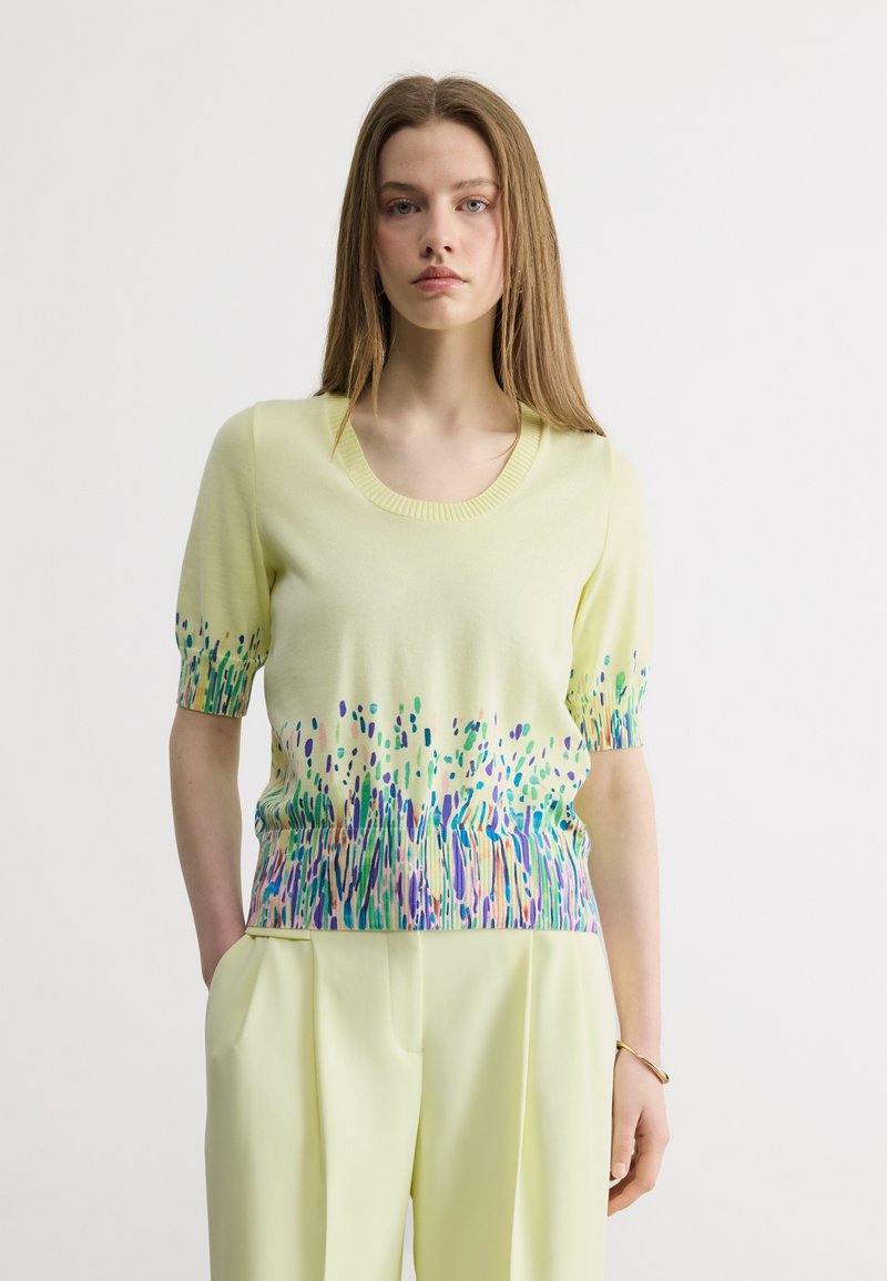 Νέα γυναίκα με κοντομάνικη μπλούζα σε απαλό κίτρινο χρώμα με πολύχρωμο αφηρημένο σχέδιο και ταιριαστό κίτρινο παντελόνι, που στέκεται με τα χέρια στις τσέπες.