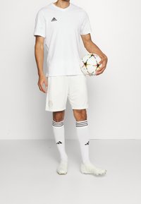 Camisa atlética blanca y pantalones cortos de Adidas, con logo negro, acompañados de calcetines blancos con rayas negras y botas de fútbol blancas, sosteniendo un balón colorido.