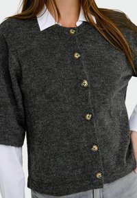 Une femme porte un cardigan en tricot gris charbon à manches courtes avec cinq gros boutons sur une chemise blanche à col et un pantalon gris clair.