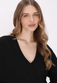 Pull noir à col en V avec des manches longues et une texture douce, porté par un mannequin aux cheveux bruns ondulés, sur un fond neutre.