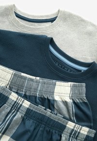 Trei topuri texturate în gri și albastru închis, cu un model tricotat, suprapuse peste șorturi de pijama în carouri, de culoare albastră și crem.