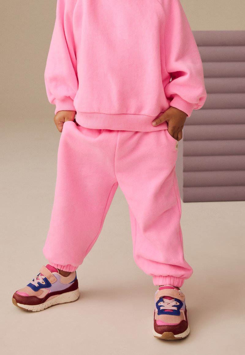 Bambino che indossa pantaloni della tuta rosa acceso, felpa abbinata e sneakers colorate con accenti blu, rosa e beige, in piedi con le mani nelle tasche.