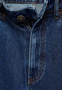 Temno modri denim hlače z zadrgo in značilnim bakrenim šivanjem. Opozarja se na teksturirano tkanino in subtilno bledečo robove.