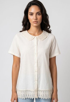 Mujer que lleva una blusa color crema de manga corta con botones, con cuello y ribete de encaje en el dobladillo, combinada con vaqueros azul claro.