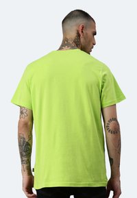 T-shirt in cotone verde lime con maniche corte, collo rotondo e vestibilità ampia. Presenta tatuaggi visibili sulle braccia.