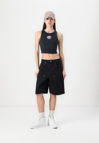 Black cropped tank top with pink logo, black knee-length shorts with front pockets, white platform sneakers, and beige cap.