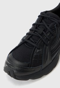Black athletic shoe with mesh upper, leather overlays, thick sole, and black laces tied in a crisscross pattern.