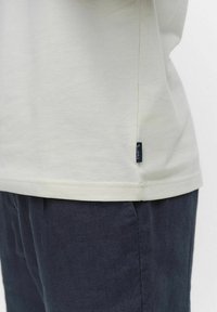 Ljust beige t-shirt i bomull med korta ärmar, rak nederkant och en liten märkestagg i marinblått på sidan, kombinerad med mörka byxor.