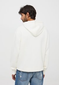 Felpa bianca con cappuccio regolabile e polsini a coste, abbinata a jeans chiari strappati; tessuto morbido con una texture liscia.