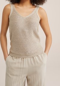 Ljust beige stickad tanktop med V-ringning och breda axelband, med en subtil glans och ribbad nederkant. Matchas med ljusbeige veckade byxor.