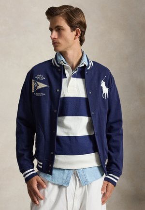 Mladić odjeven u višeslojnu tamnoplavu jaknu jedriličarskog kluba preko plavo-bijele prugaste ragbi majice i svijetloplavu chambray košulju na kopčanje.