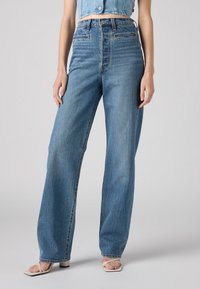 Jeans bleu clair taille haute avec une coupe droite, présentant une texture en denim délavé et des détails de couture subtils, associés à des sandales beiges.