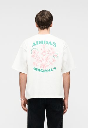 Férfi, fehér, túlméretezett Adidas Originals pólót visel rózsaszín virágmintával és zöld logós felirattal a hátán, fekete farmerrel párosítva.