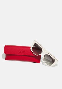 Occhiali da sole bianchi oversize con lenti grigie, caratterizzati da un motivo decorativo. Accompagnati da una custodia in pelle rossa con texture e il logo "GUESS" impresso.