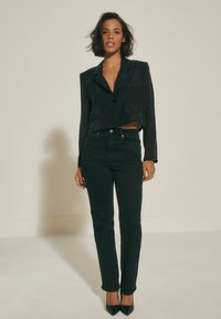 Svart kort blazer med knappstängning, tillsammans med högmidjade svarta jeans och spetsiga svarta klackar, mot en neutral bakgrund.