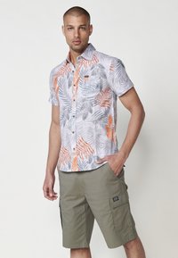 Camicia button-up a maniche corte con motivi a foglie arancioni e grigi su uno sfondo chiaro, abbinata a pantaloni cargo verde oliva con tasche.