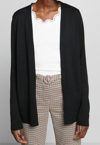 Cardigan noir à devant ouvert avec une texture côtelée, manches larges et poches latérales, porté sur un haut blanc et un pantalon à motif à carreaux.