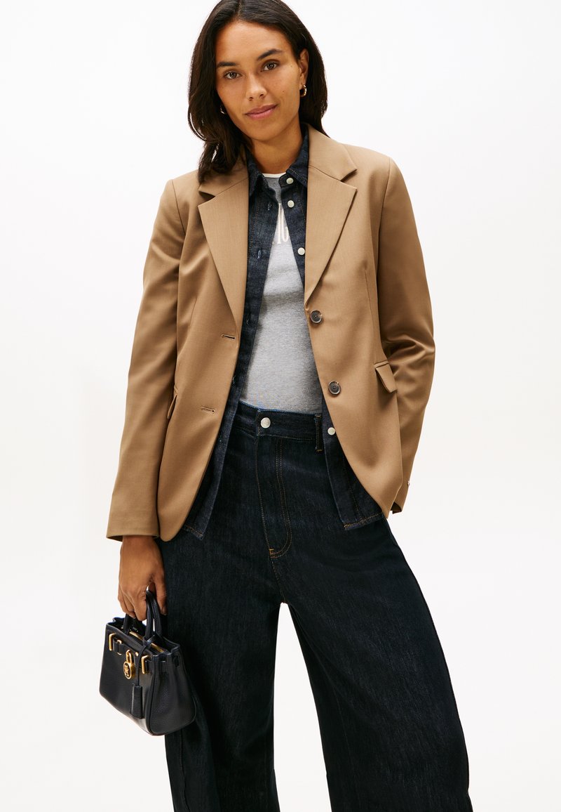 Femme portant un blazer beige sur une chemise en jean et un haut gris, jean noir à jambes larges, tenant un sac à main bleu marine sur fond blanc.