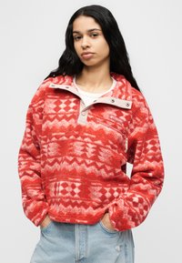 Κόκκινο φλις πουλόβερ με γεωμετρικά σχέδια, γιακά με φερμουάρ μέχρι τη μέση και αντίθετο ανοιχτό ύφασμα στον γιακά και στην τσέπη στο στήθος.