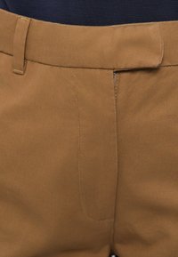 Κοντινό πλάνο καφέ παντελονιού με φερμουάρ και ζώνη, φορεμένο πάνω από σκούρο ύφασμα.