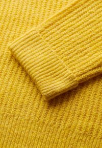 Dettaglio di un maglione giallo lavorato a maglia, con polsini a coste e motivi texturizzati su tutta la superficie, realizzato in morbido e caldo materiale di lana.