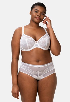 Vrouw in witte kanten bh en bijpassende slip, staand tegen een effen lichte achtergrond, met één hand haar gezicht aanrakend.