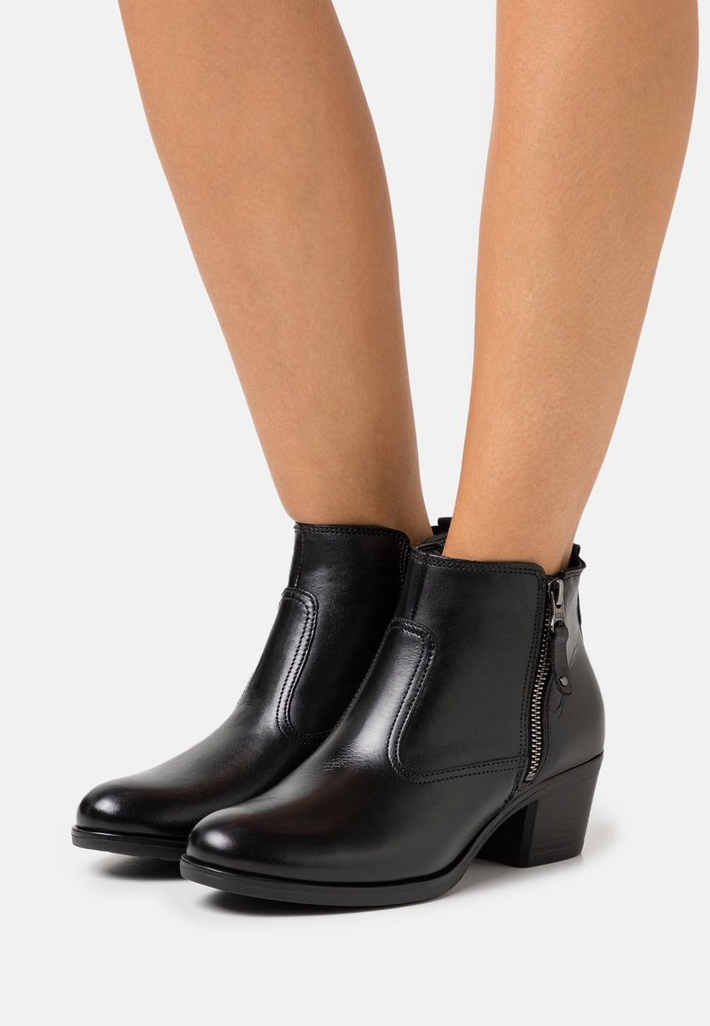 Bottines en cuir noir avec fermeture éclair latérale et petits talons carrés portées sur des jambes nues sur un fond uni.