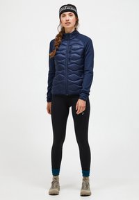 Marine blauwe geïsoleerde jas met een gestikte voorkant, getailleerd ontwerp, lange mouwen; gecombineerd met zwarte leggings en beige schoeisel. Over het geheel een gladde textuur.