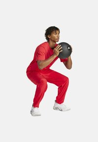 Red fitness apparel consisting of a short-sleeve shirt and pants, with a textured black medicine ball featuring a logo, in a squat position.