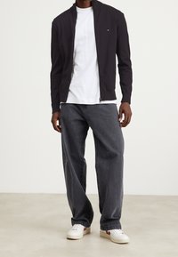 Granatowa kurtka na zamek błyskawiczny noszona na białej koszulce, w zestawieniu z luźnymi szarymi dżinsami i białymi trampkami z czerwonymi akcentami. Prosty design i luźny krój.