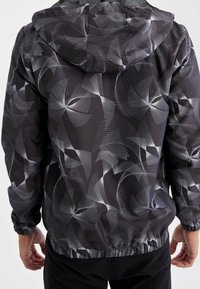 Sort hættejakke med et svulmende hvidt abstract mønster. Lavet af et glat materiale med elastiske manchetter og talje.