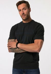 Černé tričko s krátkým rukávem vyrobené z bavlny, se sepraným designem loga. Model ho nosí s modrými džíny a černým náramkem.