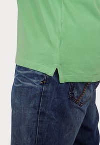 Svetlozelena polo majica z teksturirano tkanino, ovratnikom iz ravnega pletenja in stranskimi odprtinami, v kombinaciji s temno modrimi jeans hlačami z oranžnim šivom.