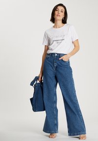 Biała koszulka z czarnym tekstem i grafiką, wysokie niebieskie dżinsowe spodnie typu wide-leg oraz niebieska torba płócienna.