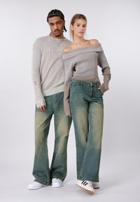 Γκρι πλεκτό πουλόβερ με ριμπ υφή, σχέδιο με πέτα στου ώμους· ξεθωριασμένο πράσινο τζιν φαρδιάς γραμμής· λευκά αθλητικά παπούτσια με μαύρες ρίγες.